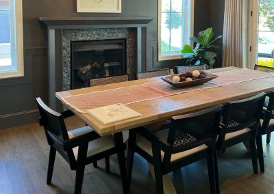 Dining room with a wooden table, six chairs, a fireplace, and a plant by the window.