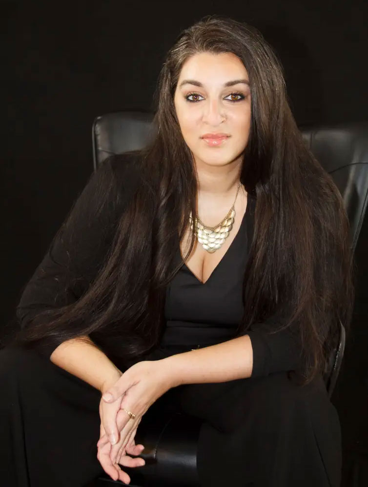 Woman with long dark hair in a black outfit, sitting on a black chair against a dark background, looking at camera.