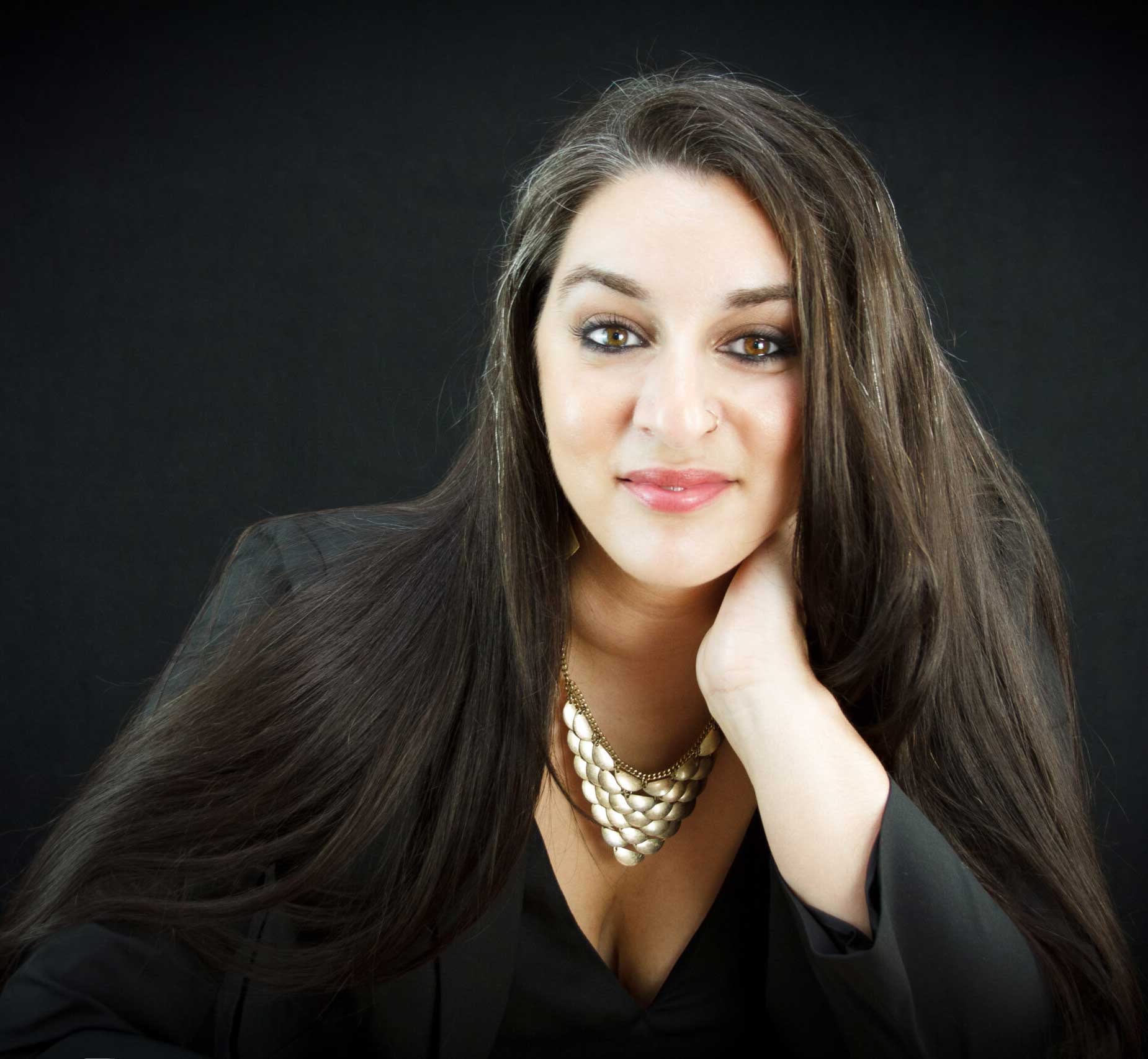 Woman with long dark hair, wearing a black blazer and gold necklace, smiling against a dark background.