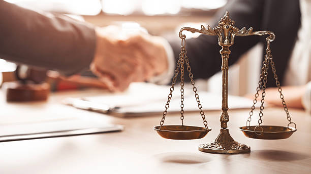 A brass balance scale on a desk with two people shaking hands in the blurred background.