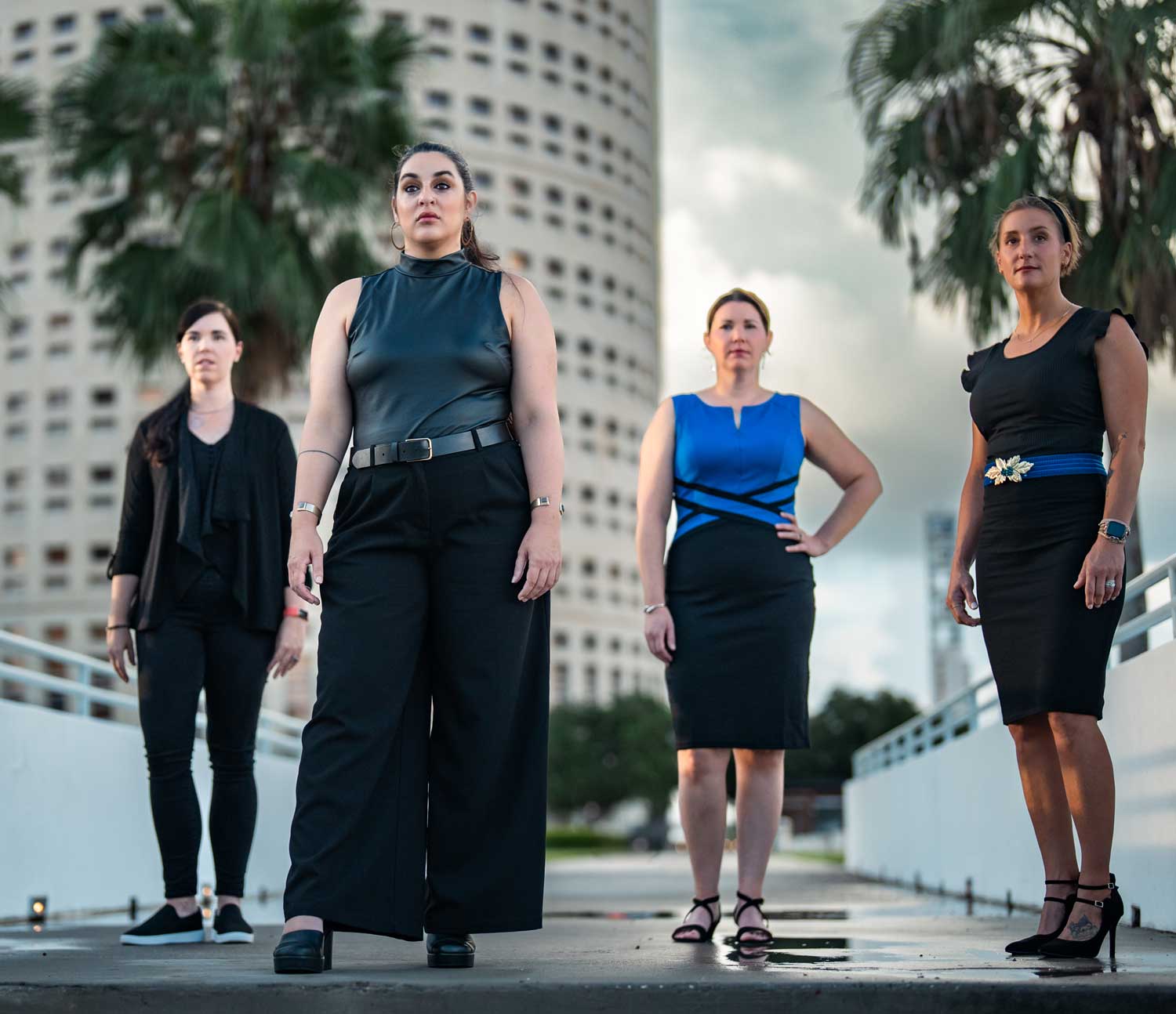 Four women in business attire stand confidently outdoors with skyscrapers and palm trees in the background.